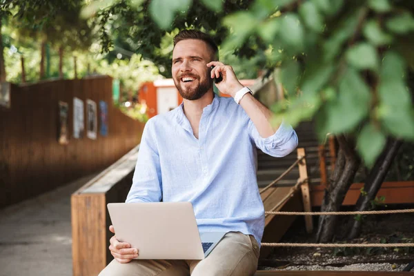 Fotoğraf gülümseyen duygusal Genç sakallı adam açık havada cep telefonuyla konuşurken dizüstü bilgisayar kullanan.