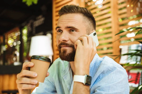 Mutlu sakallı yakışıklı genç bir kenara kahve içme cep telefonuyla konuşurken açık havada bakış fotoğraf.