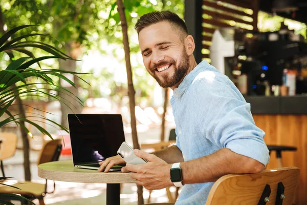 Fotoğraf gülümseyen duygusal Genç sakallı adam açık havada dizüstü bilgisayar ve cep telefonu kullanarak.