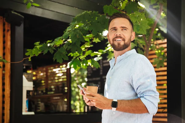 Görüntü gülümseyen Genç sakallı adam açık havada bir kenara kahve içme cep telefonu arıyorum kullanarak.