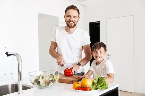 Photo of cheerful young man father dad cooking with his son indoors at home in kitchen. Looking camera.