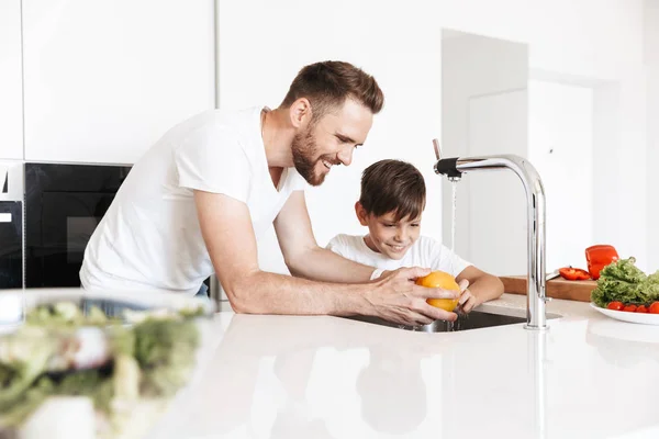 Photo of cheerful young man father dad cooking with his son indoors at home in kitchen.