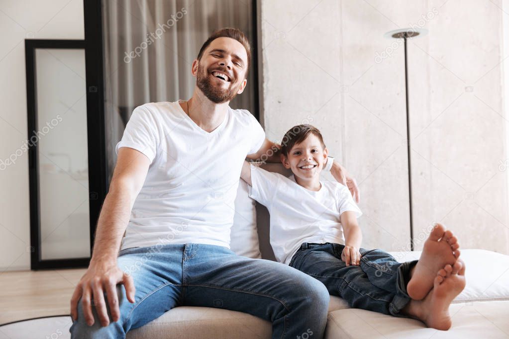 Image of cheerful young man father dad having fun with his son indoors at home watching TV.
