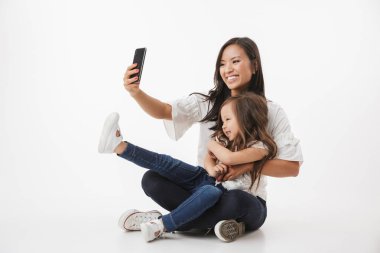 Beyaz duvar arka plan üzerinde izole görüntü bir duygusal mutlu Genç Asyalı kadın anne oturan küçük kız çocuk kızıyla birlikte yapmak selfie cep telefonuyla.