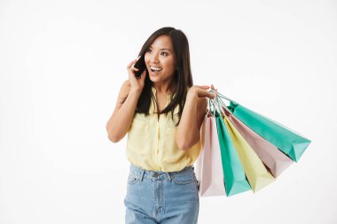 Mutlu duygusal Genç Asyalı kadın beyaz duvar arka plan üzerinde alışveriş torbaları cep telefonuyla konuşurken ile izole görüntüsünü.
