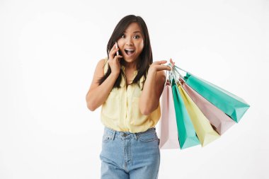 Mutlu duygusal Genç Asyalı kadın beyaz duvar arka plan üzerinde alışveriş torbaları cep telefonuyla konuşurken ile izole görüntüsünü.