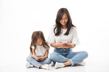 Güzel Asya Aile Anne ve küçük kızı tutan ve akıllı telefonlar ile birlikte bacaklar katta otururken kullanarak geçti, beyaz arka plan üzerinde izole