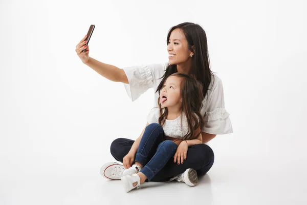 形象的情感快乐的年轻的亚洲妇女的母亲与她的小女儿的孩子坐在白色的墙壁背景下 使自拍手机 — 图库照片