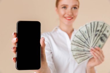 Boş ekran cep telefonu bej renkli arka plan üzerinde izole para banknot tutarken gösterilen gülümseyen bir iş kadını, yakın çekim