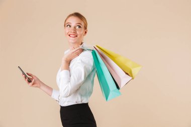 Amaç bir kenara alışveriş torbaları cep telefonuyla tutan bej duvar bakmaktan izole ayakta neşeli genç iş kadını görüntüsünü.