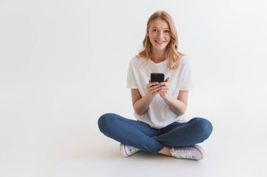 Beyaz duvar arka plan üzerinde izole oturan genç ve güzel şirin kızıl saçlı kadın resmi. Cep telefonu kullaranarak görünümlü kamera.