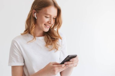 Kulaklık ile müzik dinleme beyaz duvar arka plan üzerinde cep telefonu tarafından izole duygusal sevimli genç Kızıl saçlı kadın fotoğrafı.