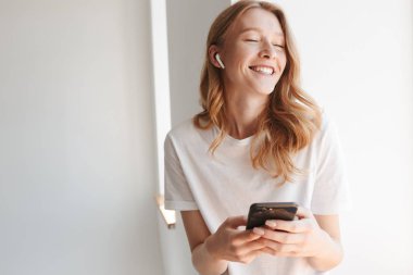 Kulaklık ile müzik dinleme beyaz duvar arka plan üzerinde cep telefonu tarafından izole genç ve güzel kızıl saçlı kadın resim.