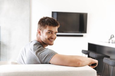 Tv Uzaktan kumanda ile evde bir kanepede oturan ve kameraya bakarak gülümseyen genç adam
