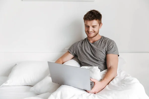 自宅のベッドに座りながらノート パソコンを使用して若い男の笑みを浮かべてください — ストック写真