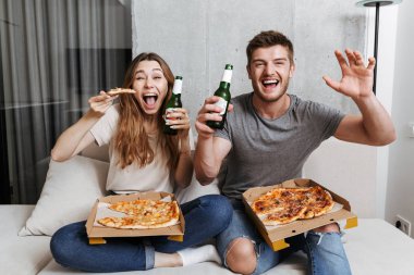 Pizza yiyip birlikte evde kanepede oturan ve Tv izlerken bira içmeyi genç çift heyecanlı