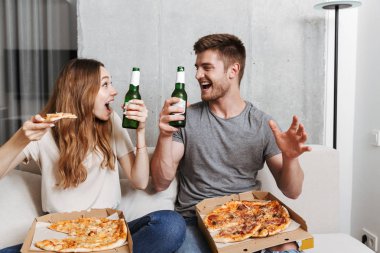 Pizza yiyip birlikte evde kanepede oturan ve Tv izlerken bira içmeyi neşeli genç çift
