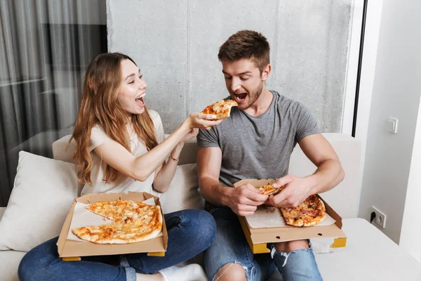 Smiling young couple eating pizza while sitting together on couch at home