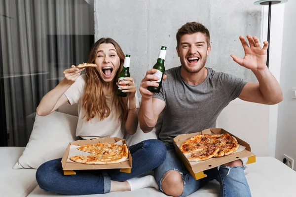 Pizza yiyip birlikte evde kanepede oturan ve Tv izlerken bira içmeyi genç çift heyecanlı