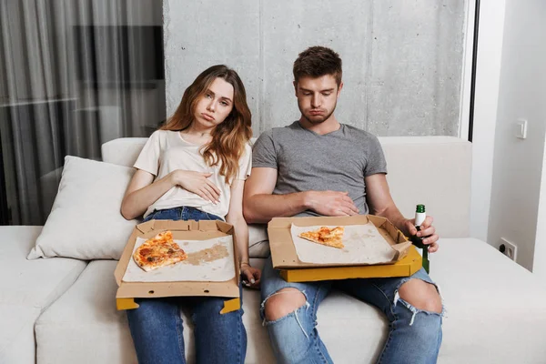 Unhappy couple ate too much pizza while sitting together on a couch at home