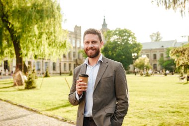 Çim ve ağaçları ile parkta yürüyüş sırasında paket servisi olan restoran kahve yudumlarken kameraya gülümseyen gri klasik elbiseli olgun neşeli adam fotoğrafı