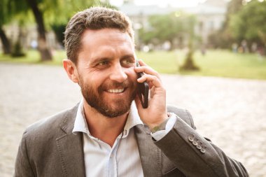 Görüntü gri Klasik takım Smartphone'da parkta yürüyüş sırasında güneşli günde konuşma içinde mutlu işadamı