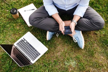 Beyaz adamın cep telefonu ve gümüş laptop kullanırken çim parkta oturan iş giyim üstten kırpılmış fotoğraf