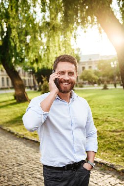 Fotoğrafı neşeli adam şehir parkı güneşli günde yürüyüş ve cep telefonu konuşma 30s