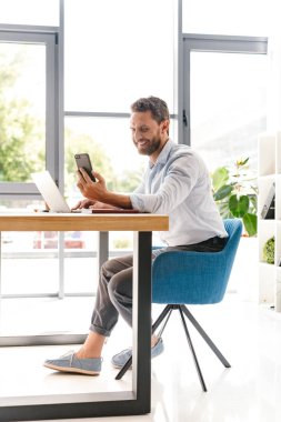 Office çalışma bir kenara cep telefonu ve dizüstü bilgisayar kullanarak seyir içinde oturan mutlu yakışıklı sakallı adam görüntüsünü.