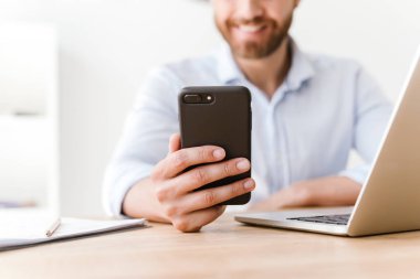 Cep telefonu ve dizüstü bilgisayar kullanarak office çalışma içinde oturan neşeli sakallı adam kırpılmış fotoğrafı.