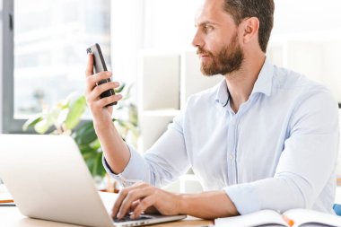 Office çalışma bir kenara dizüstü bilgisayar kullanan cep telefonuyla konuşurken seyir içinde oturan mutlu yakışıklı sakallı adam görüntüsünü.