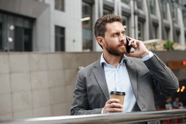 灰色西装的成熟男子照片看着一边站着 拿着外卖咖啡在现代商务中心前的商务电话 — 图库照片