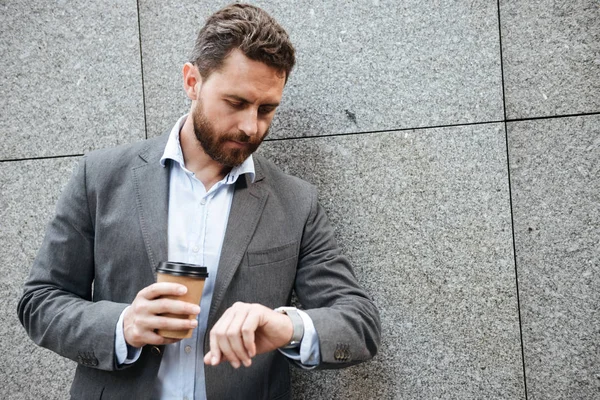 グレーのスーツで白シャツは花崗岩の壁に立っている テイクアウトのコーヒーを飲みながら腕時計を見て大人のビジネスマンのクローズ アップ写真 — ストック写真