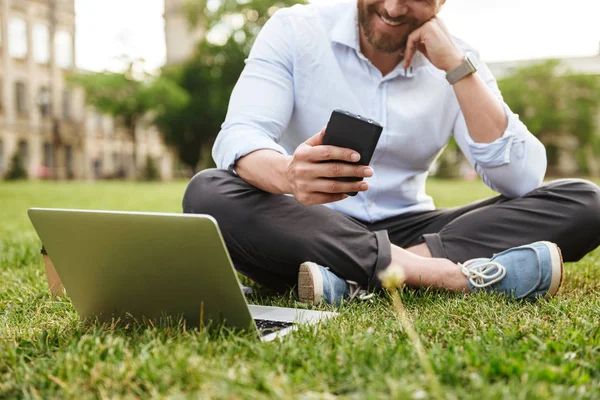 Cep telefonu ve gümüş laptop kullanırken bağdaş ile çim parkta oturan iş giyim gülümseyen adam beyaz fotoğraf kırpılmış