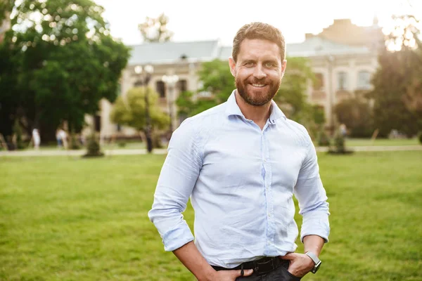 Elleri ceplerinde eski binanın arka plan üzerinde yeşil parkta yürüyüş sırasında beyaz gömlek ayakta Avrupa çekici adam portresi