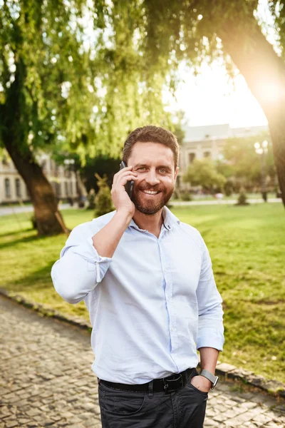 Fotoğrafı neşeli adam şehir parkı güneşli günde yürüyüş ve cep telefonu konuşma 30s