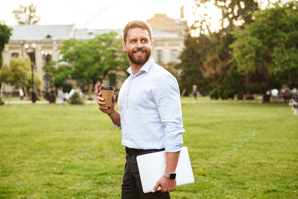 Image of successful business man in white shirt walking through park carrying takeaway coffee and silver laptop