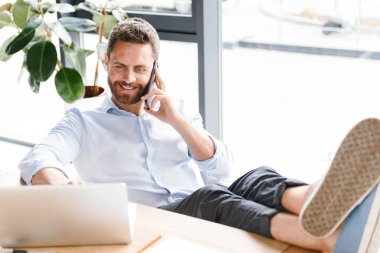 Office çalışma bir kenara dizüstü bilgisayar kullanırken cep telefonuyla konuşan bakan yakışıklı sakallı adam görüntüsünü.