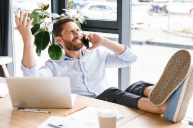 Office çalışma bir kenara dizüstü bilgisayar kullanırken cep telefonuyla konuşan bakan yakışıklı sakallı adam görüntüsünü.