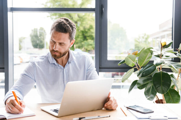 Concentrated businessman working on laptop computer while sitting at the desk in office and making notes