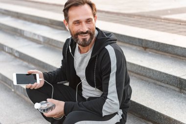 Su şişesi açık havada adımlarla üzerinde otururken cep telefonuyla kulaklık sporcu gülümseyen