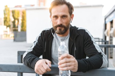 Yakışıklı güçlü olgun sporcu şişe su ile tutarak kulaklık ile müzik dinleme açık havada görünümlü kamera oturan görüntüsünü.