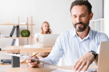 Bir arka plan ile işbirliği işçisi kadın ofiste otururken dizüstü bilgisayar ve belgeler üzerinde çalışan gülümseyen adam Yöneticisi