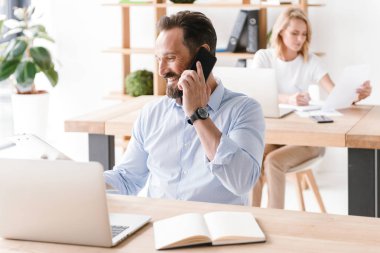 Mutlu bir adam Yöneticisi ofiste oturan ve bir arka plan üzerinde işbirliği işçisi kadın ile cep telefonu üzerinde konuşurken dizüstü bilgisayar ve belgeler ile çalışma