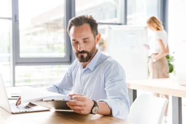 Bir arka plan ile işbirliği işçisi kadın ofiste otururken dizüstü bilgisayar ve belgeler üzerinde çalışan odaklı adam Yöneticisi