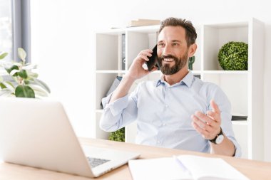 Evrak ve dizüstü bilgisayar ile ofis masa başında otururken cep telefonu üzerinde konuşurken heyecan iş adamı