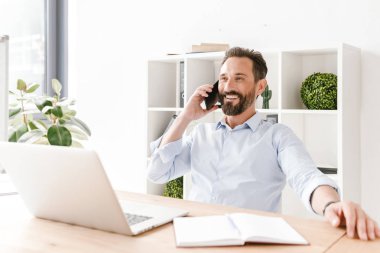 Evrak ve dizüstü bilgisayar ile ofis masa başında otururken cep telefonu üzerinde konuşurken gülümseyen iş adamı