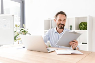 Heyecanlı işadamı klasör evrak ve dizüstü bilgisayar ile ofis masa başında otururken holding