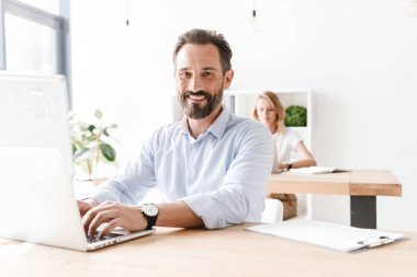 Mutlu bir adam Yöneticisi bir arka plan ile işbirliği işçisi kadın ofiste otururken dizüstü bilgisayar ve belgeler üzerinde çalışma