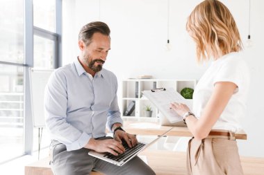 Adam Yöneticisi holding laptop evrak ile ayakta işbirliği işçisi kadın ile ofiste bir masa üzerine eğilerek süre gülümseyen